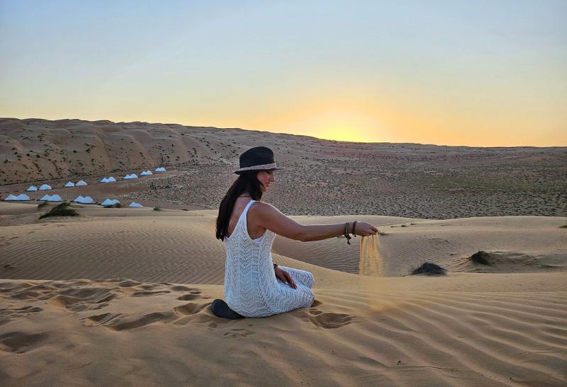 Tmavovlasá žena v béžových šatách na dunách v Ománe.