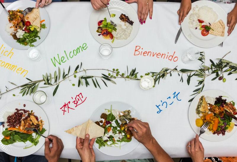 Stôl s nápismi "vitajte" v rôznych jazykoch, jedlo na tenieroch a ľudia sediaci za stolom.