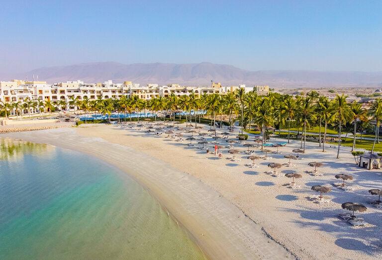 Piesková pláž, palmy kokosové, lehátka a budova hotela Fanar v Ománe.