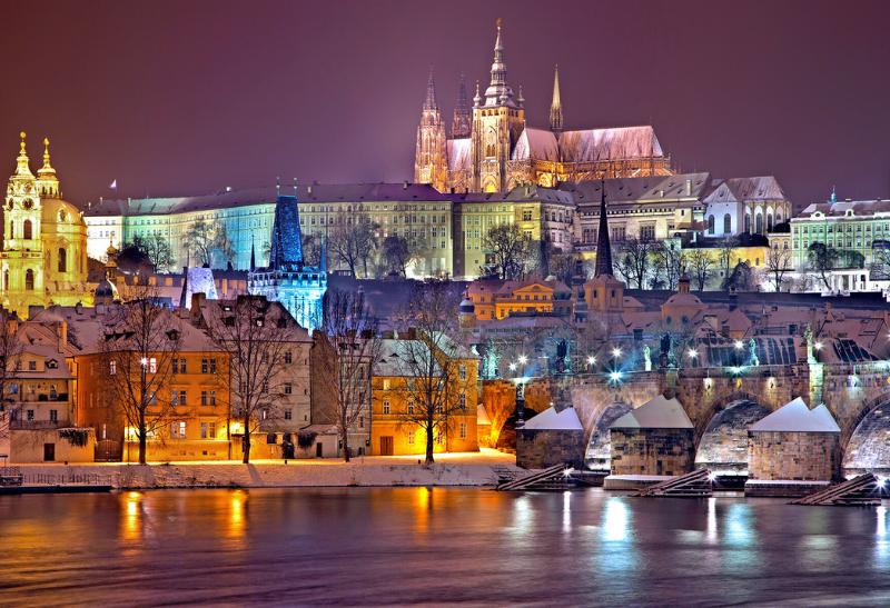 Vysvietená Praha s hradom, budovami, riekou a so snehovou pokrývkou.
