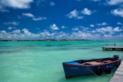 Modrá loď priviazaná o drevené mólo, tyrkysové more a zelený ostrov v pozadí. Maurícius