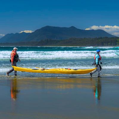 Piesočnatá pláž, oceán a zeleň pri pobreží a ľudia nesúci kajak.