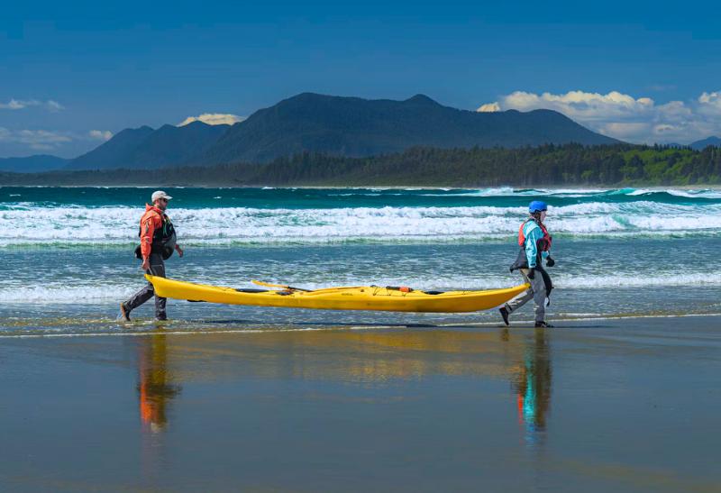 Piesočnatá pláž, oceán a zeleň pri pobreží a ľudia nesúci kajak.